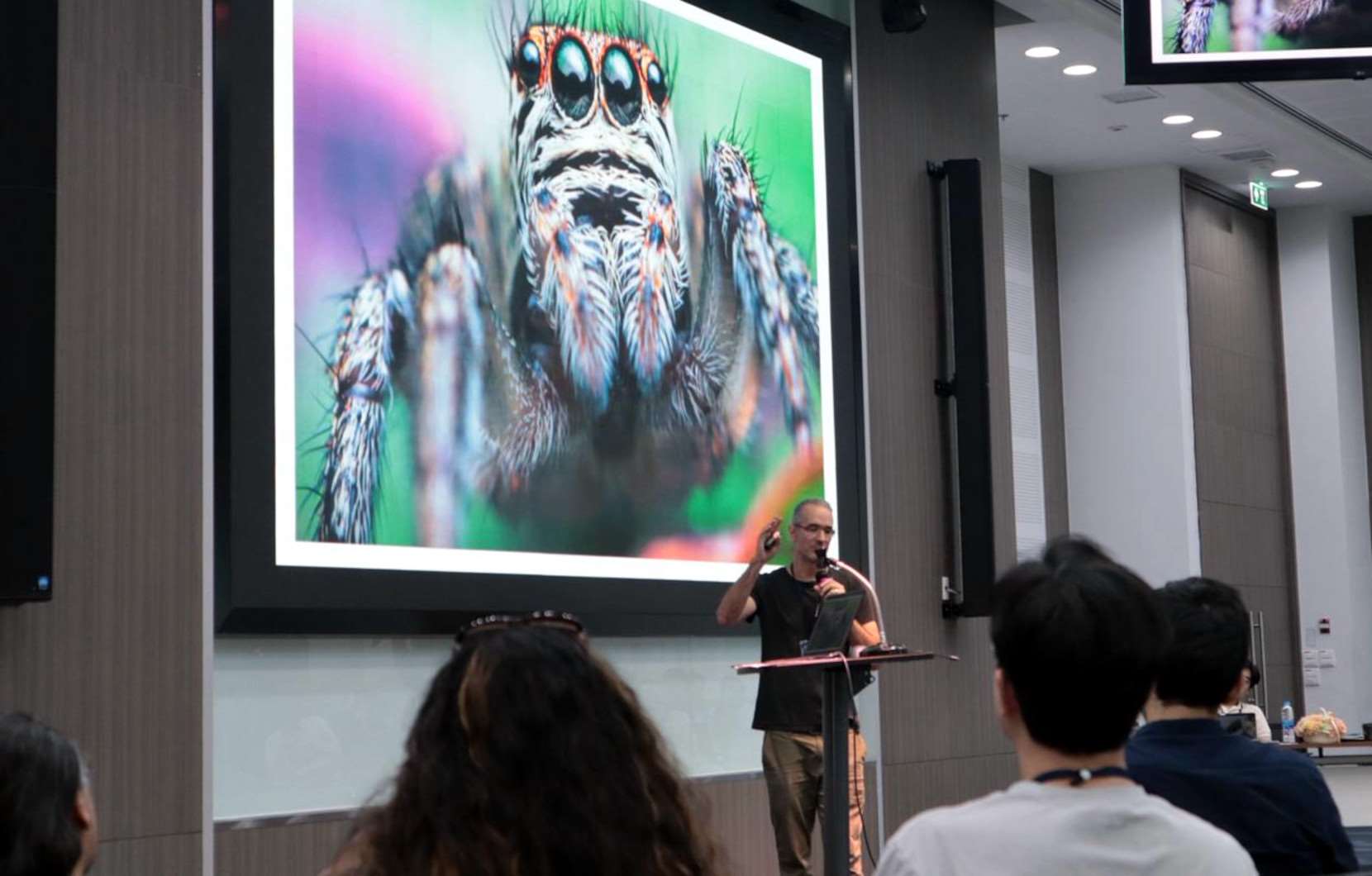 Prof. Christoph Dahl bei einem Vortrag auf einer wissenschaftlichen Konferenz. Im Hintergrund ist eine Springspinne zu sehen, die als Modellorganismus in seiner Forschung zu Tierverhalten, Kognition und Wahrnehmung eine wichtige Rolle spielt. (Foto: Mit freundlicher Genehmigung von Prof. Christoph Dahl)