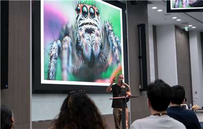 Prof. Christoph Dahl bei einem Vortrag auf einer wissenschaftlichen Konferenz. Im Hintergrund ist eine Springspinne zu sehen, die als Modellorganismus in seiner Forschung zu Tierverhalten, Kognition und Wahrnehmung eine wichtige Rolle spielt. (Foto: Mit freundlicher Genehmigung von Prof. Christoph Dahl)