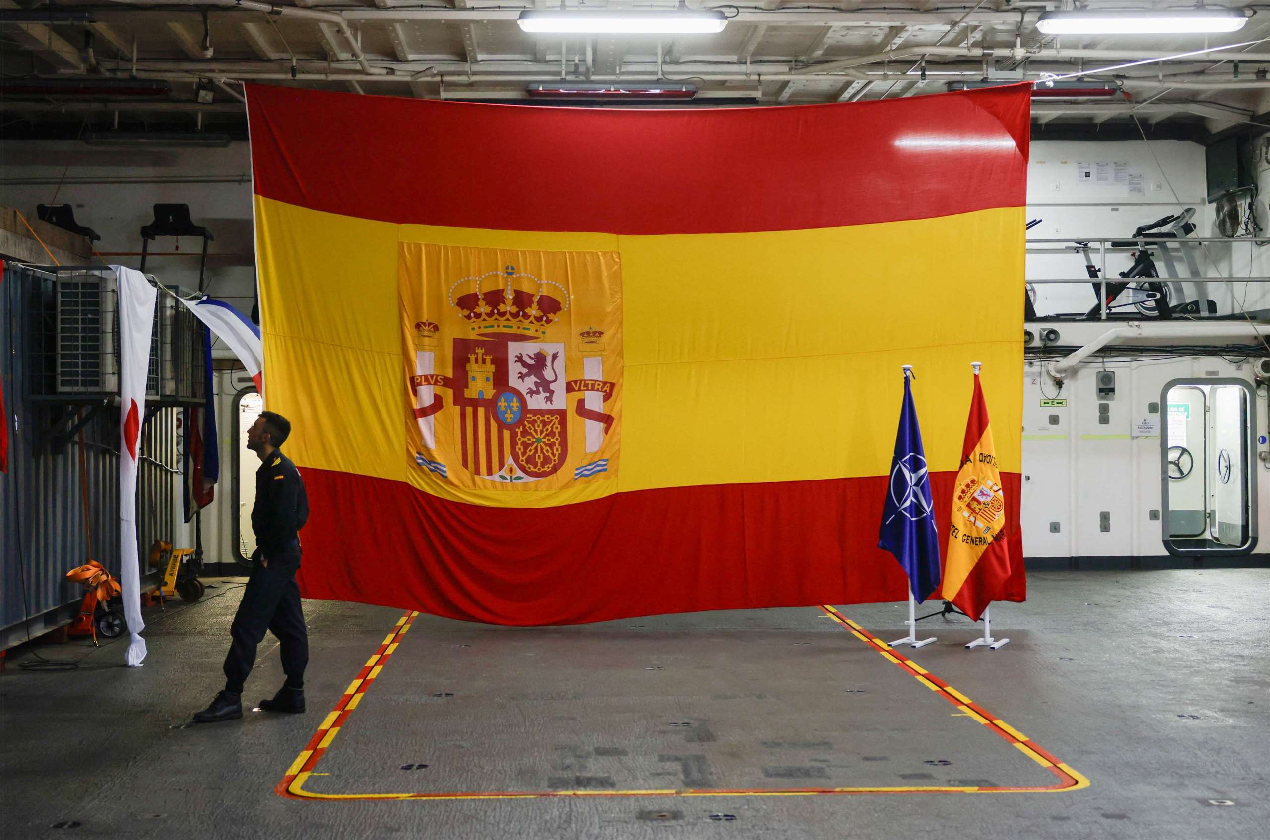 西班牙尚未達成對北約承諾的軍費目標。圖為西班牙海軍士兵站在兩棲登陸艦卡斯蒂利亞號的機庫內，身旁豎立著西班牙國旗與北約旗幟。