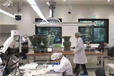 圖為中國三星堆文物保護與修復館，桌上器物從左至右為青銅大面具、青銅尊與頂尊跪坐人像。(翻攝維基百科)