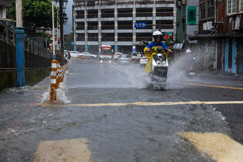 氣象署26日持續發布大規模豪雨預報，提醒西半部仍要嚴防致災性的降雨，尤其是南台灣和北台灣。圖為新北市淡水區因大雨造成路面有積淹水情形，騎士經過激起水花。